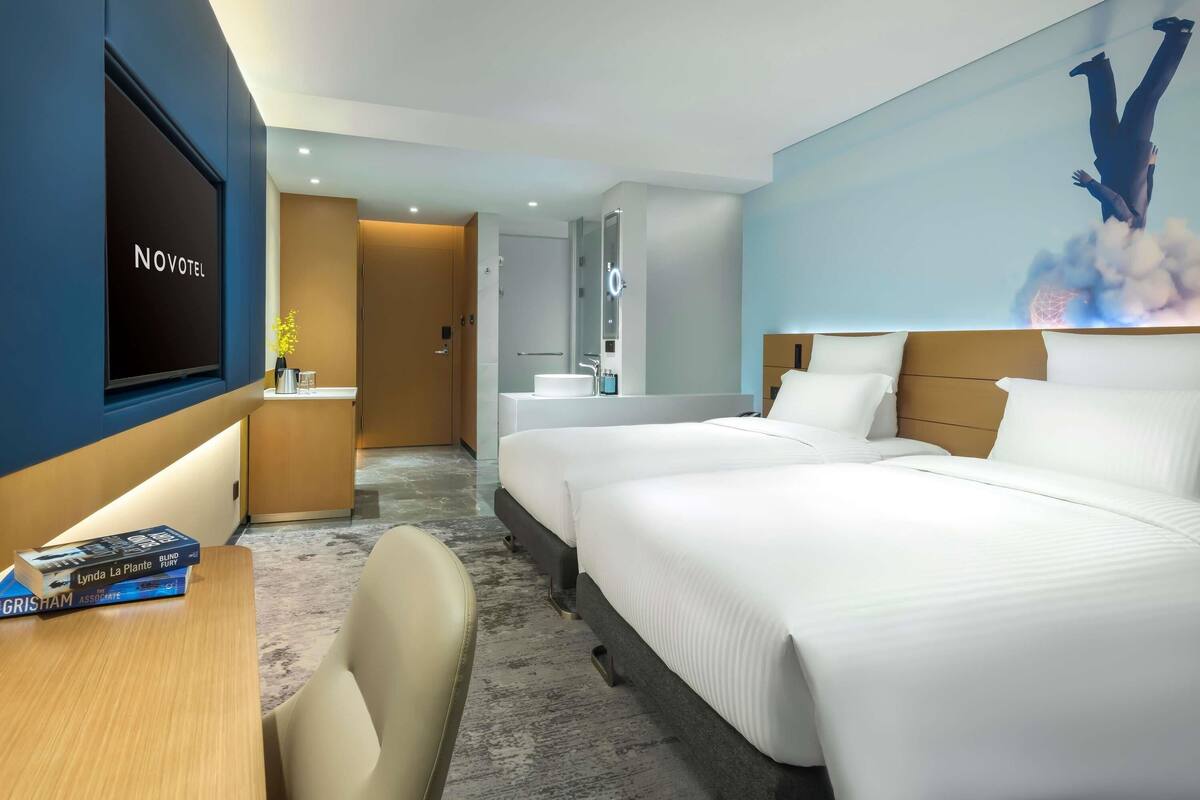 A hotel room with two beds, a TV, a desk with books, a bathroom, and a wall mural.
