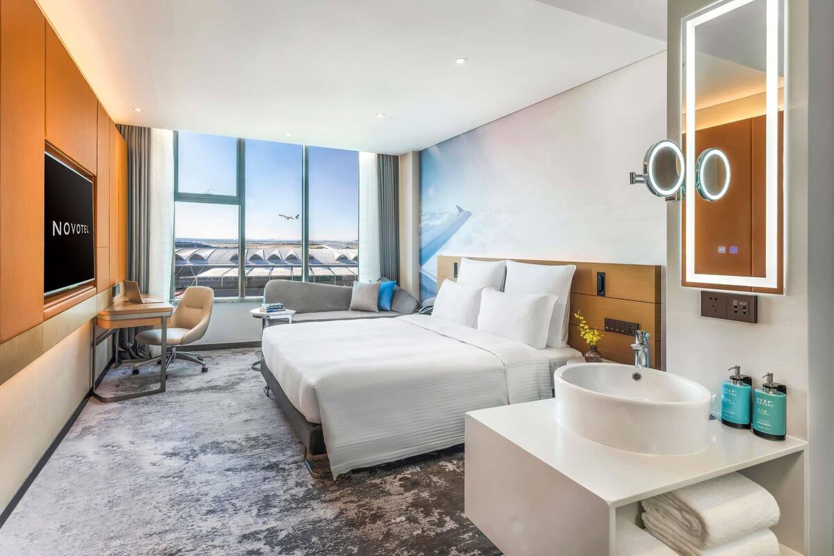 A hotel room with a bed, a desk, a chair, a large window with a view of an airport, and a bathroom with a sink and mirror.