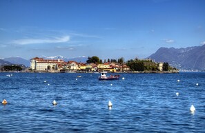 A waterfront cityscape with a prominent building complex, a boat on the water, and mountains in the background. - Peaceful Apartment with balcony, Garden And Lake View (Stresa)