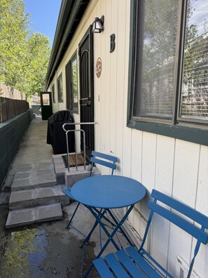 Ein überdachter Außenbereich mit Sitzgelegenheit, einem Tisch, Stühlen, einem Grill und einer Tür zu einer Laube.
