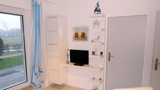 Una habitación con una televisión sobre un mueble blanco, una puerta y una ventana con vista a un jardín.