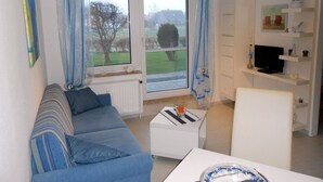 Una sala de estar con un sofá azul, una mesa de centro blanca y un televisor sobre una estantería.