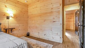 A wooden-paneled room with a bed, a small table, a lamp, and a door leading to another room.