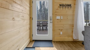 A wooden cabin interior with a door leading to an outdoor area, a welcome sign, and a bench outside.