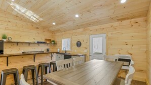 A wooden-paneled room with a long dining table, bar stools, and a kitchen area.