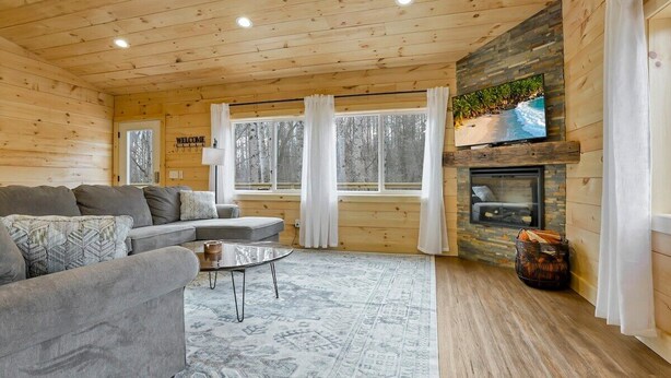 A cozy cabin interior with a wooden ceiling, a fireplace, a sofa, and a television mounted above it.