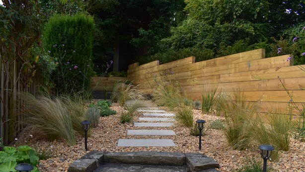 Un sentier de jardin bordé de pierres plates, entouré de différentes plantes et d’un mur en bois.