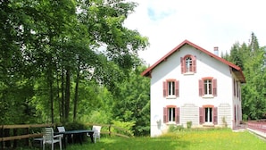 A white house with red shutters, surrounded by a wooden fence and a grassy area with a table and chairs. - L'ETERLOU in a charming property in the heart of the Source-Bleue area (Montperreux)