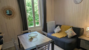 A room with a dining table, chairs, a sofa, a clock, and a window with curtains. - L'ETERLOU in a charming property in the heart of the Source-Bleue area (Montperreux)