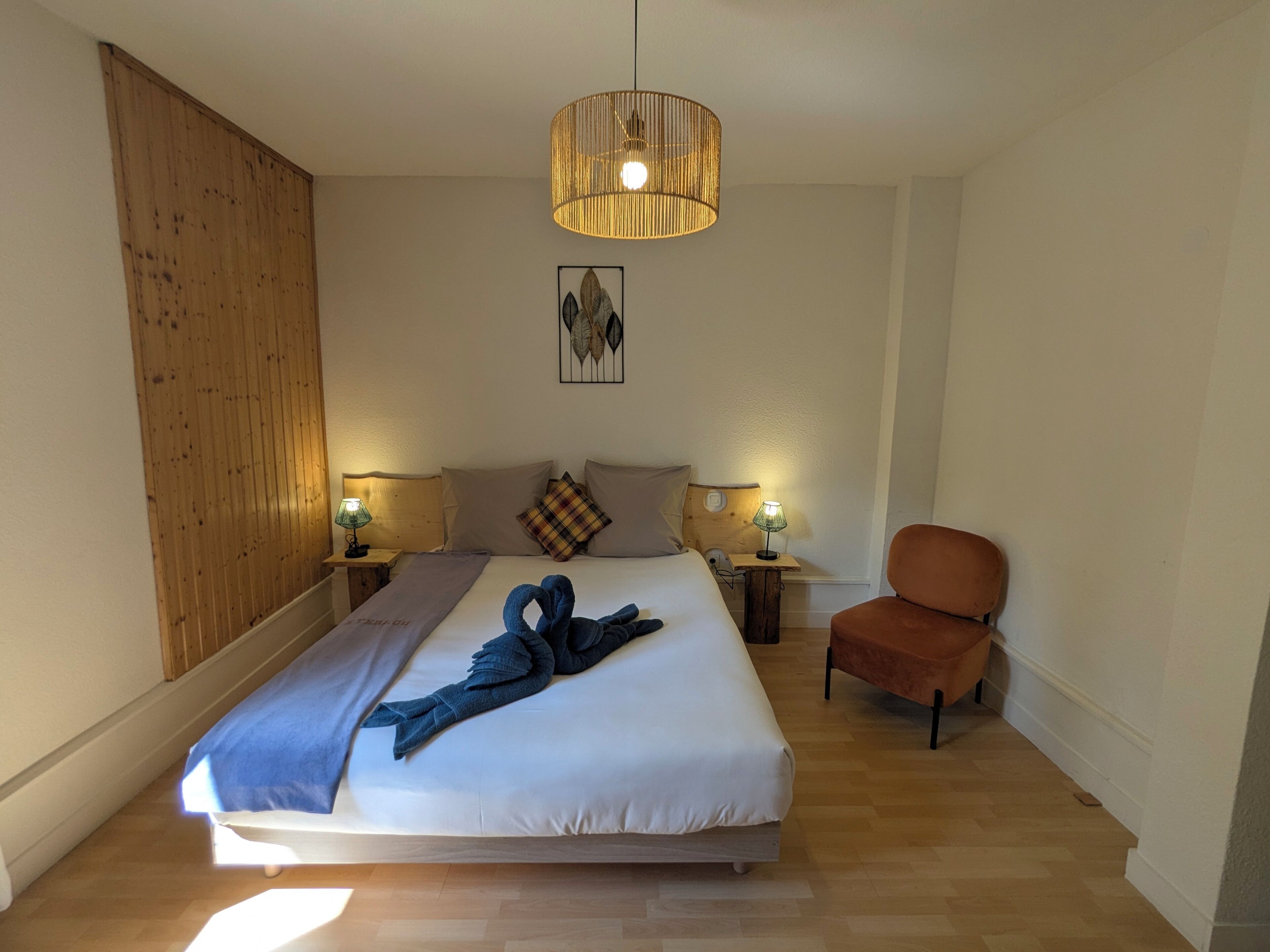 Habitación de hotel con cama, dos lámparas de noche, cabecero de madera, una obra de arte en la pared y un sillón marrón.