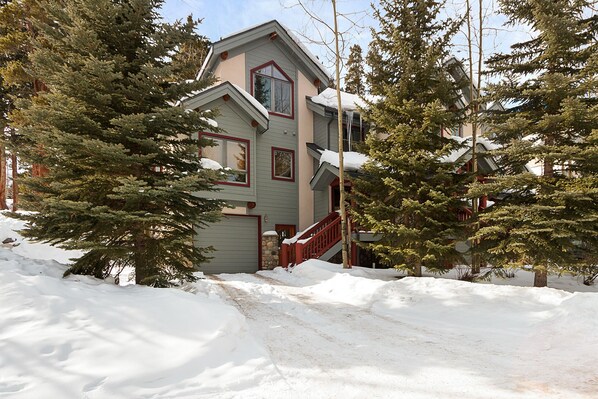 A two-story house with a stone foundation, a red front door, and a snow-covered driveway.