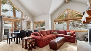 Un salón espacioso con un amplio sofá seccional rojo, una mesa de centro de madera y un comedor con mesa y sillas. Amplias ventanas ofrecen vistas del exterior.