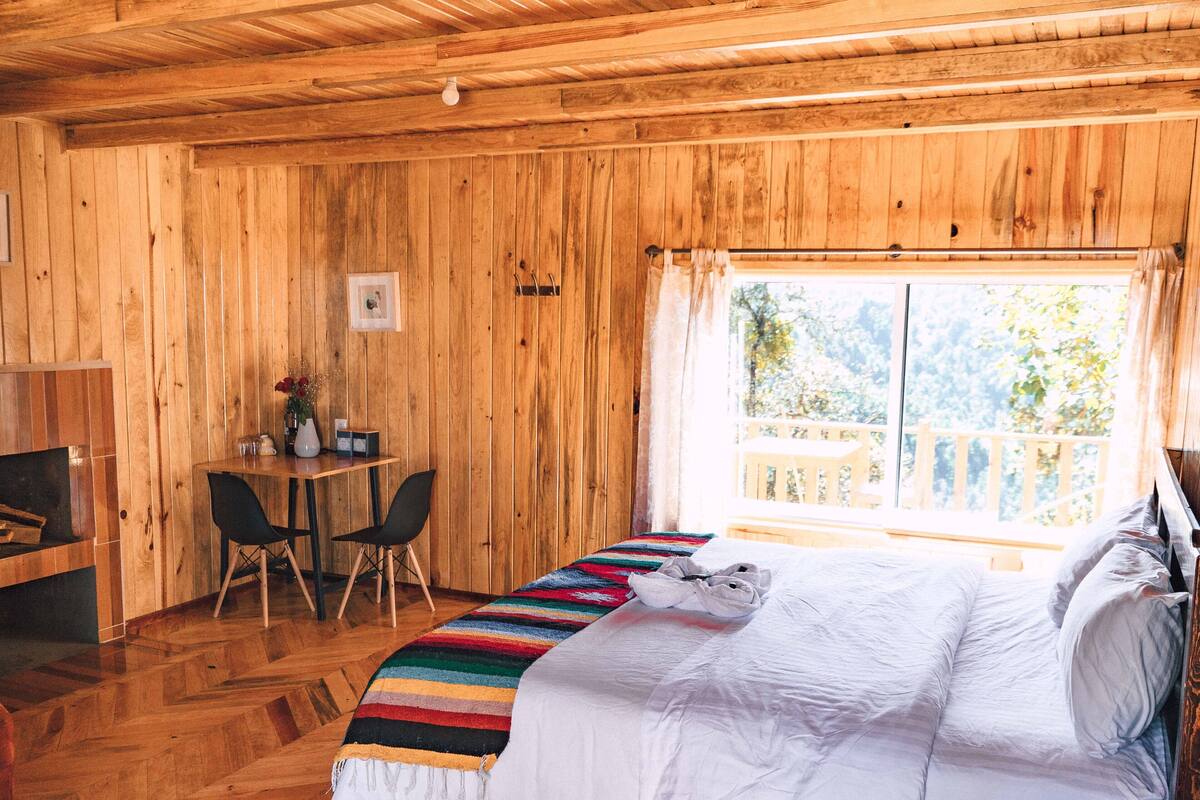 Une pièce de cabane en bois comprenant un lit, une table avec des chaises et une fenêtre donnant sur la forêt.