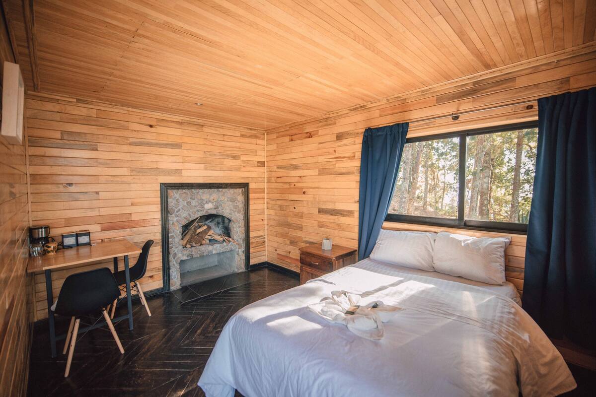 Une pièce de cabane en bois comprenant un lit, une petite table, des chaises, une cheminée et une fenêtre avec des rideaux.
