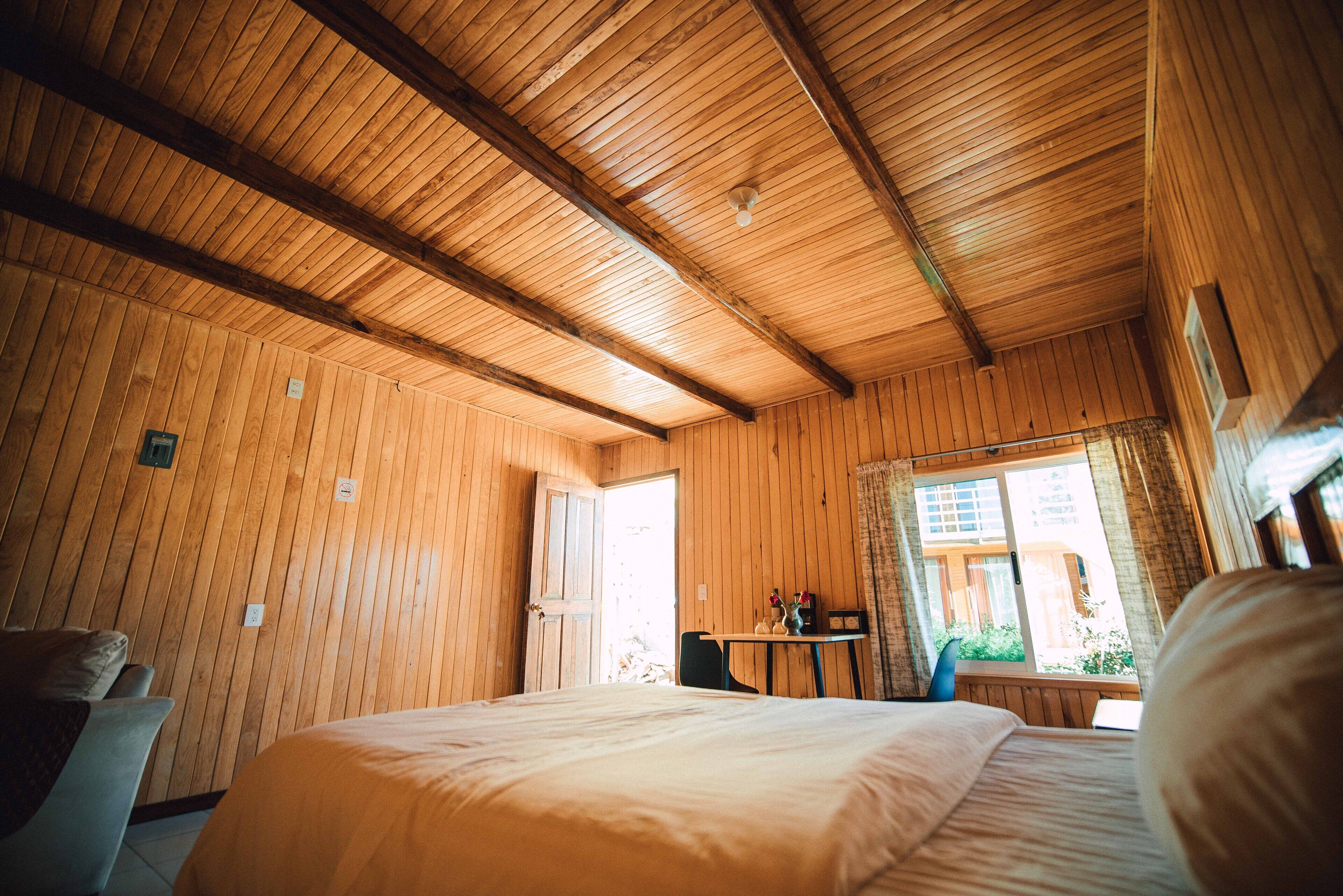 A wooden-paneled room with a bed, a table with a vase, and a window with a view of greenery.
