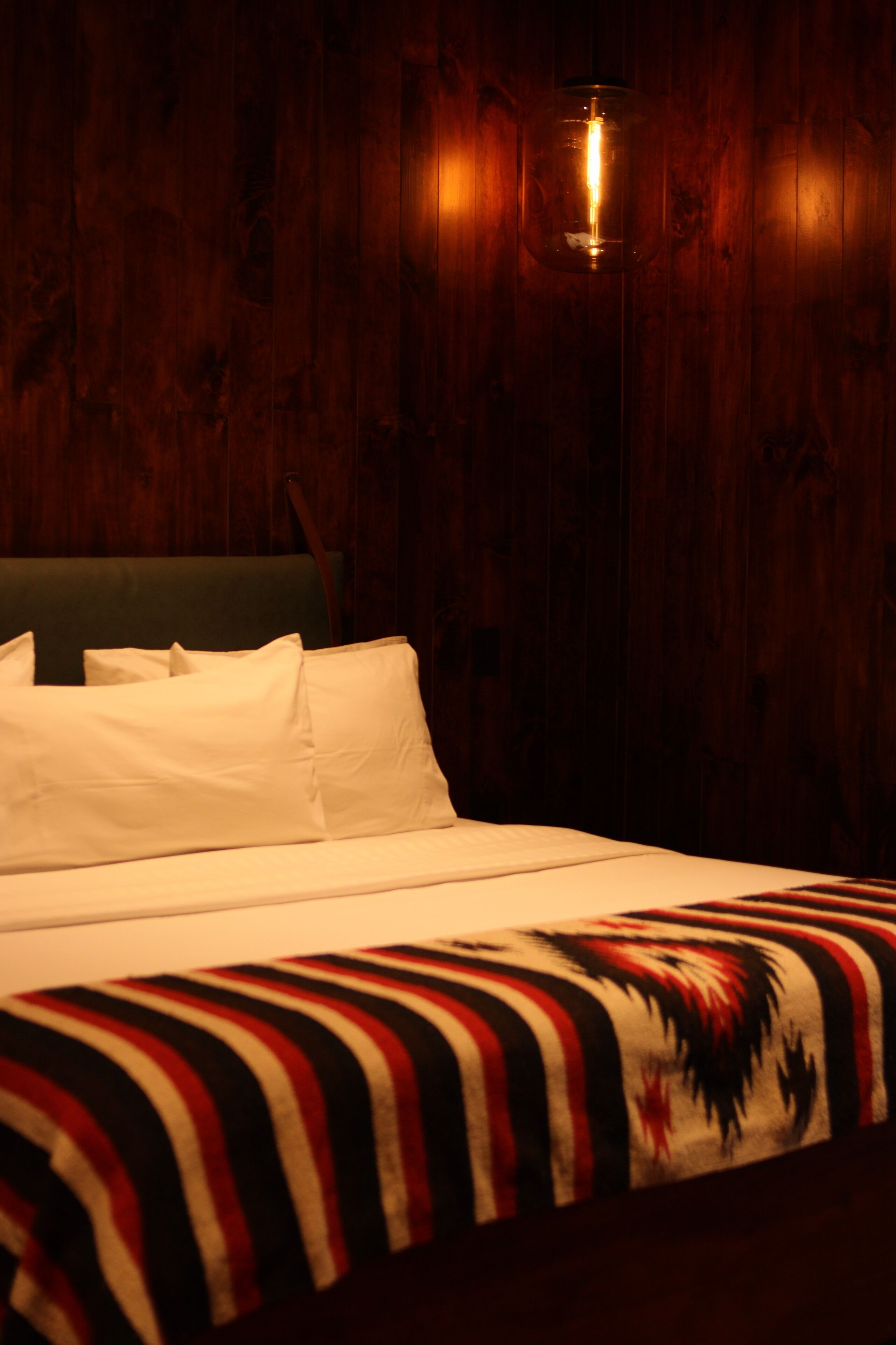 A bed with white bedding and a patterned blanket, set against a wooden wall with a hanging light fixture.