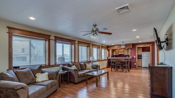 A spacious living room with a large brown sofa, a coffee table, and a ceiling fan. There is a kitchen area with wooden cabinets and a dining table. - Riverview Townhouse I in Downtown Alexandria Bay (Alexandria Bay)