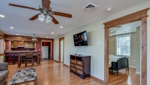 A spacious living area with hardwood flooring, a ceiling fan, a flat-screen TV, and a kitchen in the background. - Riverview Townhouse I in Downtown Alexandria Bay (Alexandria Bay)