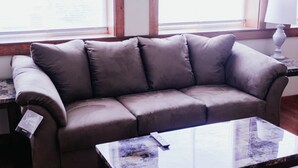 A gray sofa with a remote control on a glass coffee table in a room with wooden floors and a window with blinds. - Riverview Townhouse I in Downtown Alexandria Bay (Alexandria Bay)