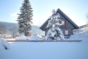 A wooden cabin with a sloped roof, surrounded by snow-covered trees and a clear sky.