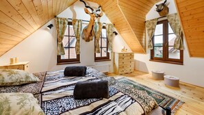 A high-ceilinged attic bedroom with wooden beams, a bed with patterned bedding, two windows with curtains, and a mounted animal head.