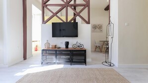 A modern living room with a large flat-screen TV mounted on the wall, a wooden console table with decorative items, a woven rug, and a dining area with a table and chairs in the background.