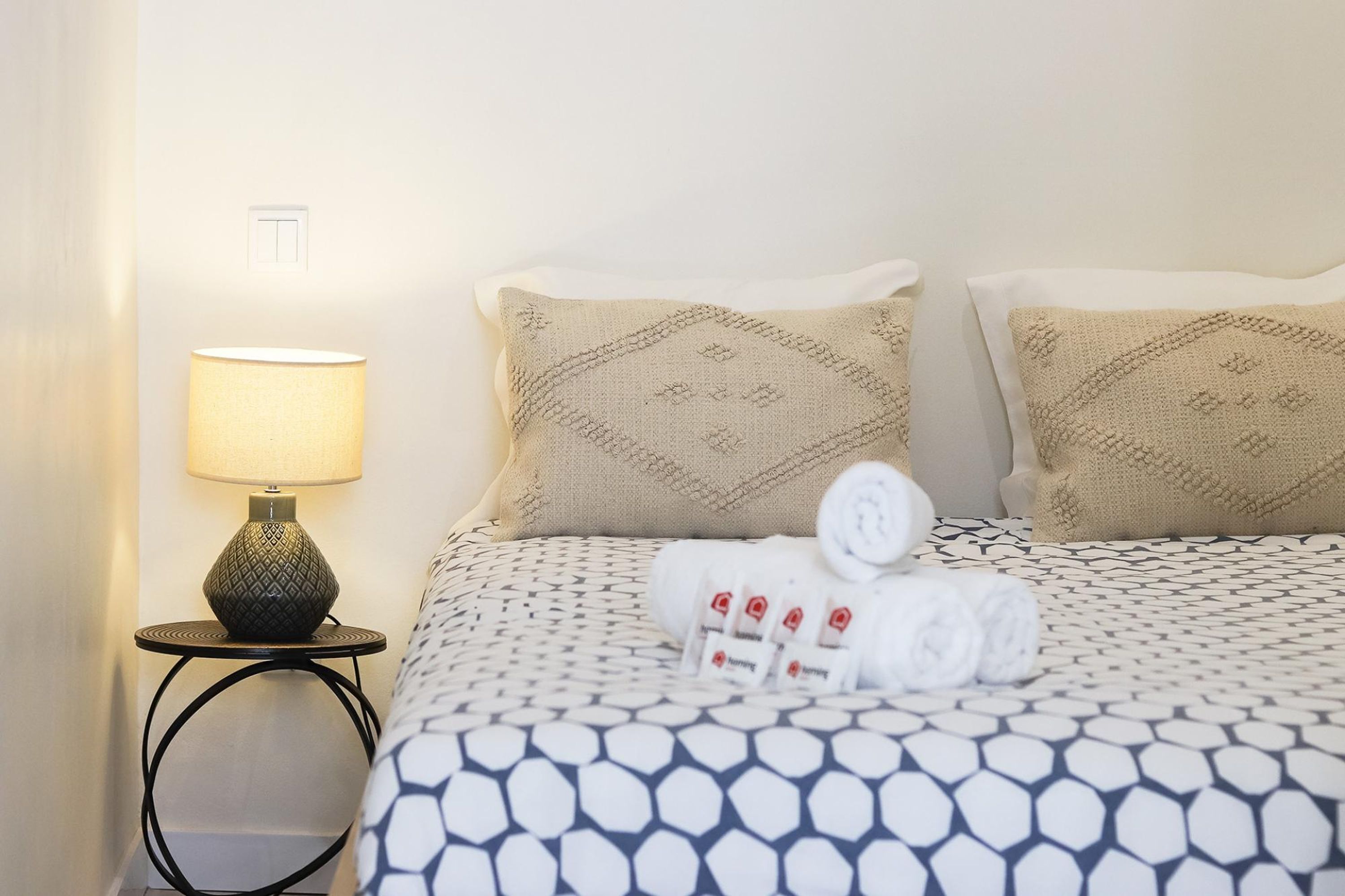 Una cama bien hecha con ropa de cama estampada, una lámpara de noche y toallas enrolladas sobre la mesita de noche.