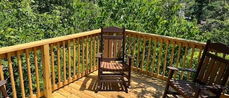 Une terrasse en bois, agrémentée de deux chaises et d’une table, également en bois, et entourée d’une balustrade en bois, d’où l’on peut admirer une zone boisée.