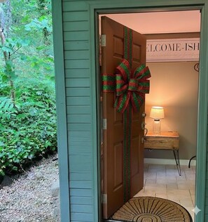 A wooden door with a large bow decoration, a welcome sign, a small table with a lamp, and a chair. - Whiskey Ridge Lodge ~ Rustic Luxe Cabin with Hot Tub, Game Lounge & Epic Mountain Views (Gatlinburg)
