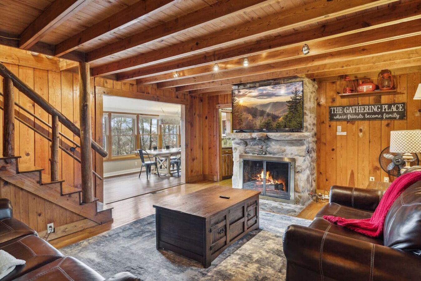 A cozy log cabin interior with a stone fireplace, wooden floors, leather furniture, and a dining area with large windows.