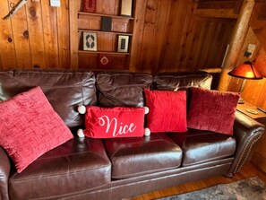 A brown leather sofa with three red pillows, one with the word 'Nice' on it, in a room with wooden walls and a lamp.