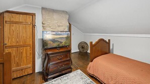Une chambre avec une armoire en bois, une commode, un lit recouvert d’une couverture orange, un téléviseur posé sur un meuble en bois et un mur en pierre.