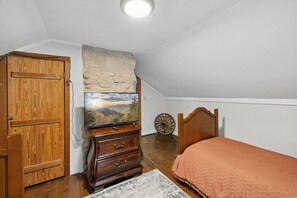 Ein Schlafzimmer mit Holzschrank, Schminktisch, Bett mit orangefarbener Decke, Fernseher auf Holzständer und Steinwand.