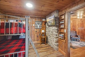 A cozy cabin room with a bunk bed, a stone wall, a fireplace, a bookshelf, and a leather armchair.