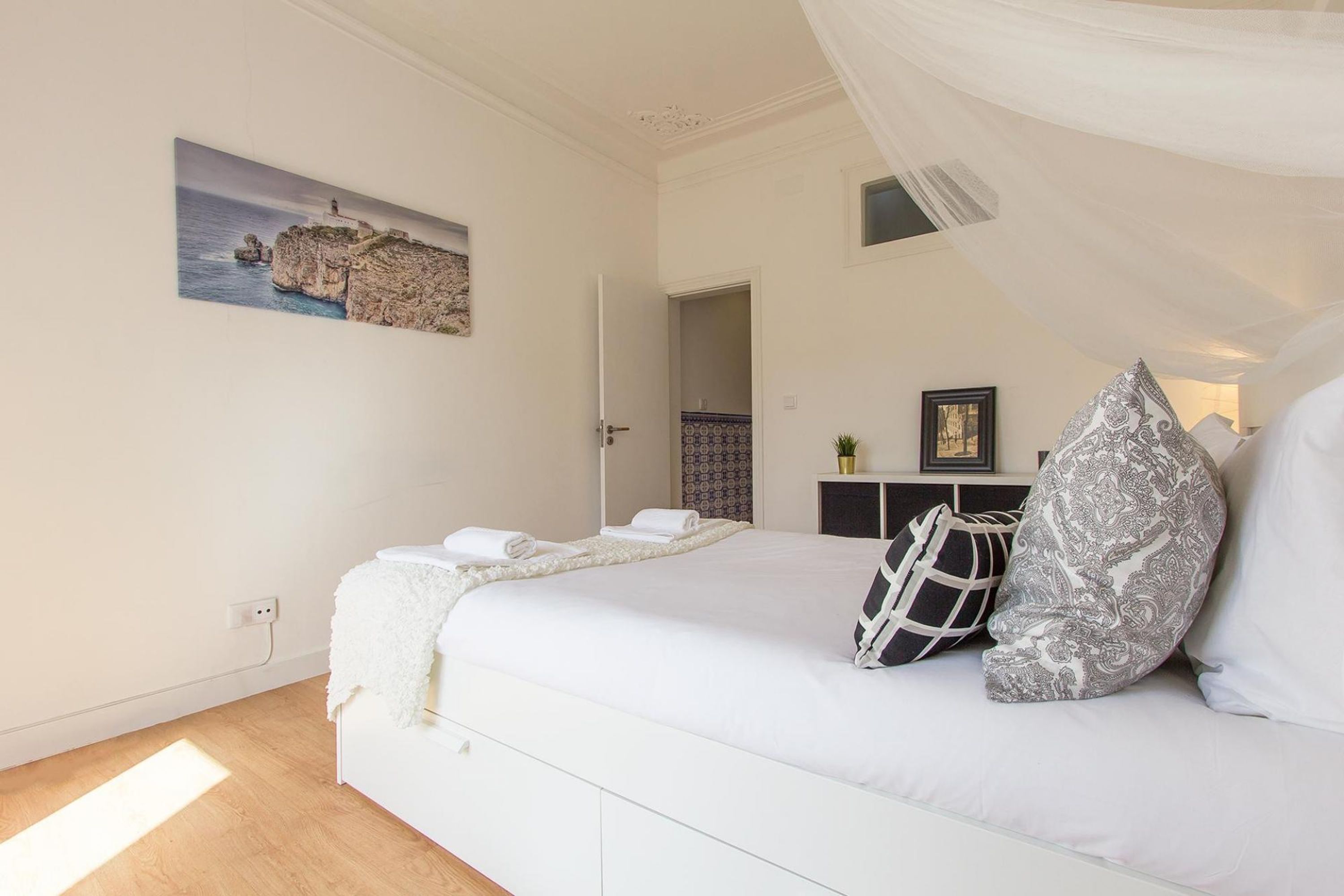 A bedroom with a bed, a painting of a coastal scene, a bedside table with a lamp, and a window with curtains.