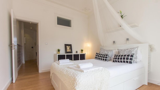 A bedroom with a white bed, a bedside table, a nightstand, a window, and a door. - Bairro Alto Stylish by Homing (Lisbon)