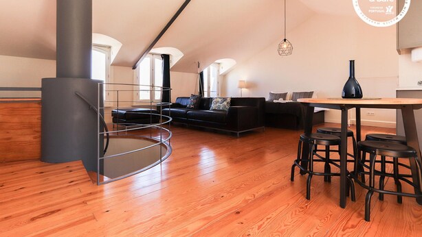 Amplio espacio tipo loft con techo alto, escalera circular, sofá de cuero negro, mesa de comedor con taburetes y lámpara colgante.