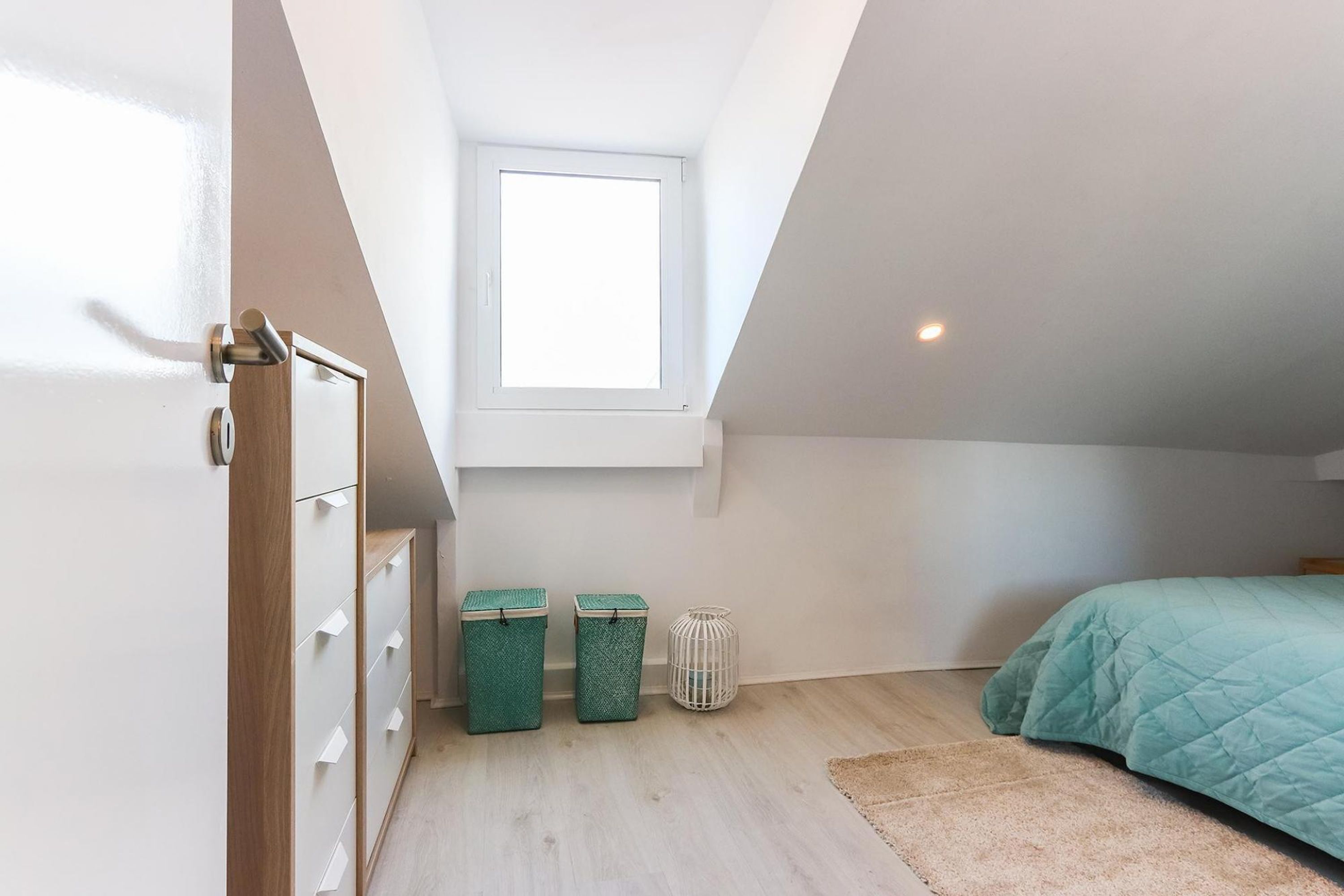 Une chambre avec un plafond en pente, une fenêtre, un lit recouvert d’une courtepointe turquoise, une armoire en bois et deux poubelles.
