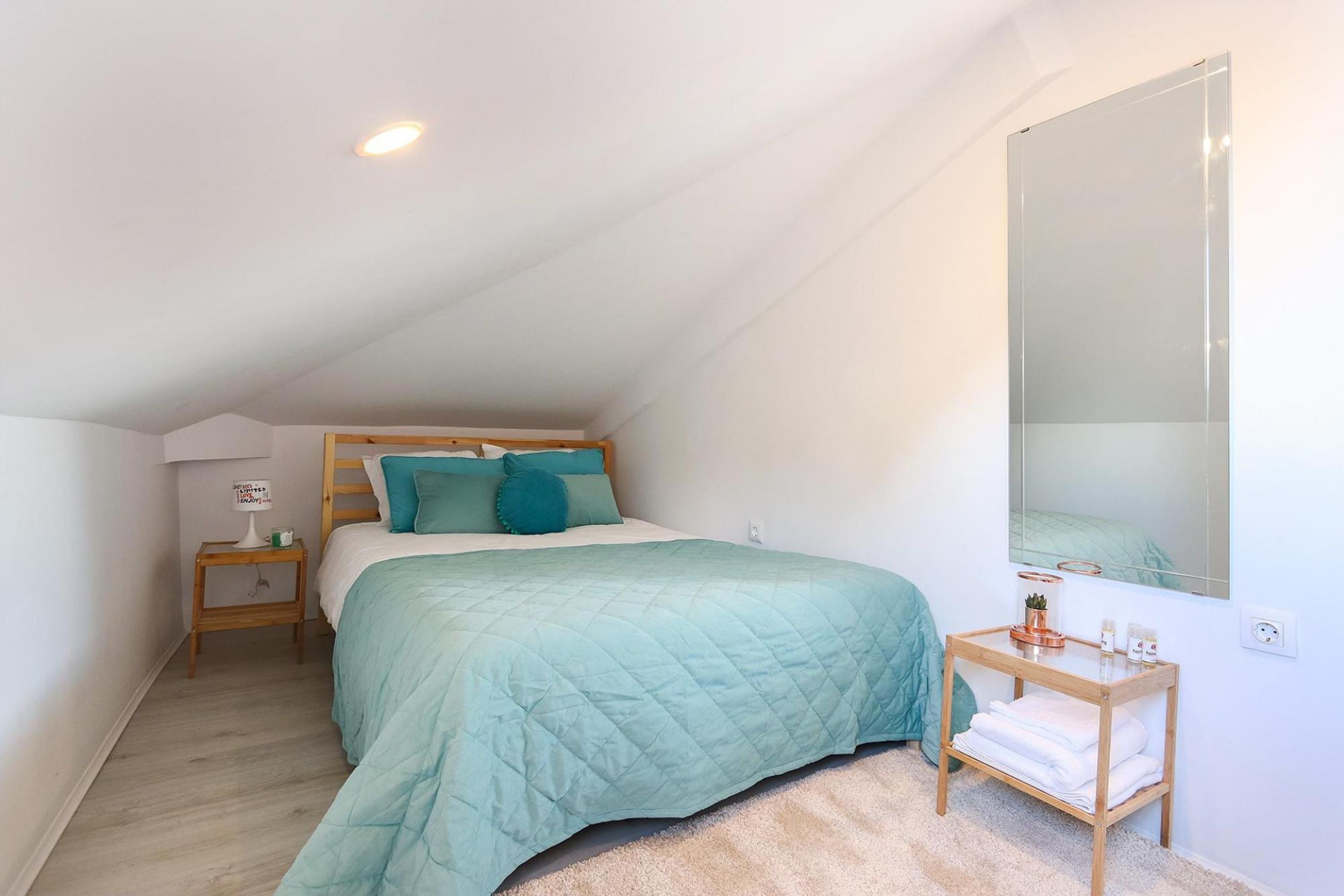 A bedroom with a bed, a nightstand, a bedside table, a mirror, and a small wooden table with towels.