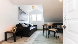 Een compacte leefruimte met een zwarte bank, een kleine tafel en een stoel. Een schilderij hangt aan de muur en er is een raam met natuurlijk licht.