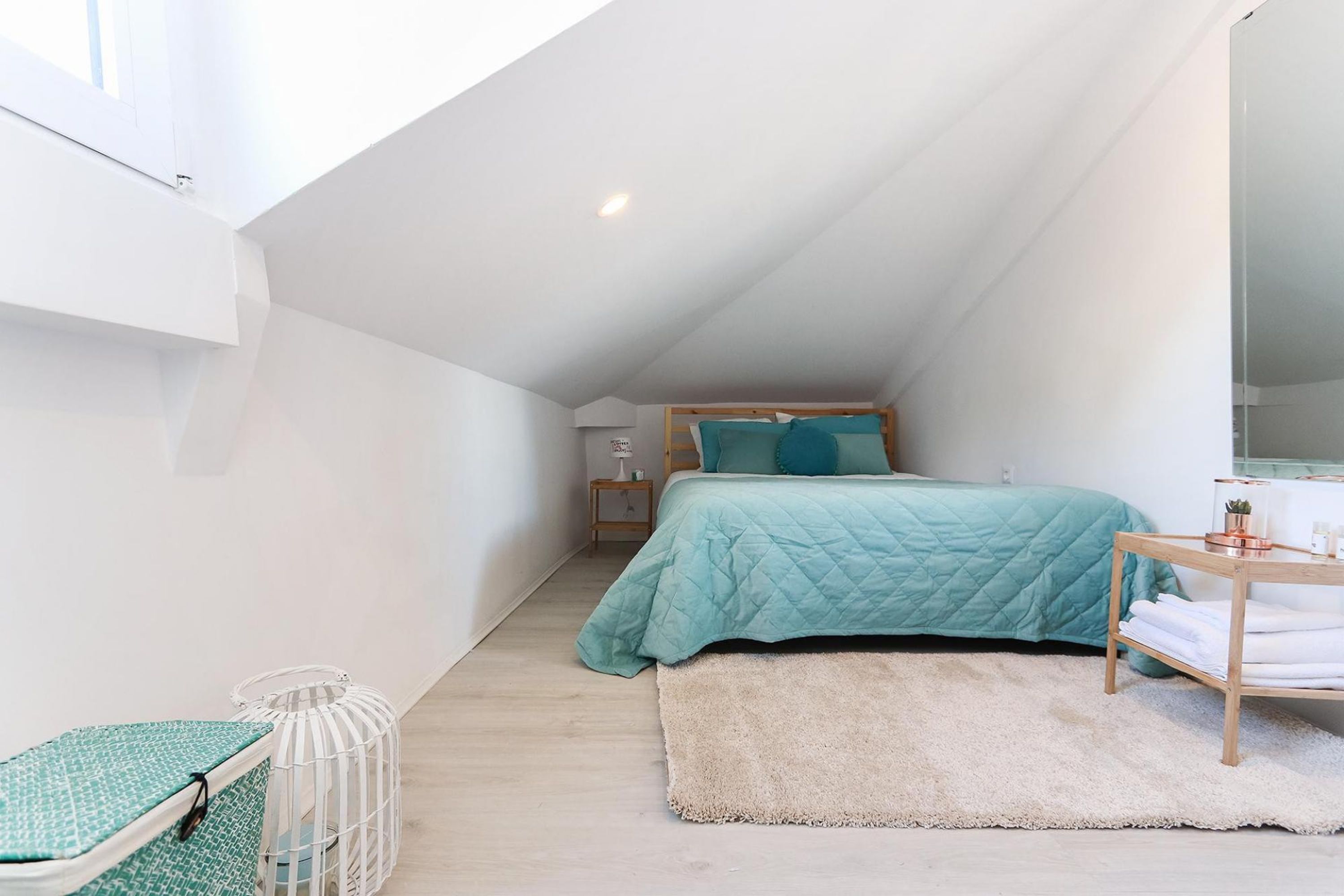 Ein kleines, gut beleuchtetes Schlafzimmer im Dachgeschoss mit schrägen Wänden, einem Bett mit türkisfarbenem Bettzeug, einem Nachttisch mit Lampe und einem kleinen Fenster.