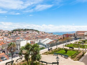A panoramic view of a historic city with a prominent hill, a waterfront, and a well-maintained public square with benches and greenery.