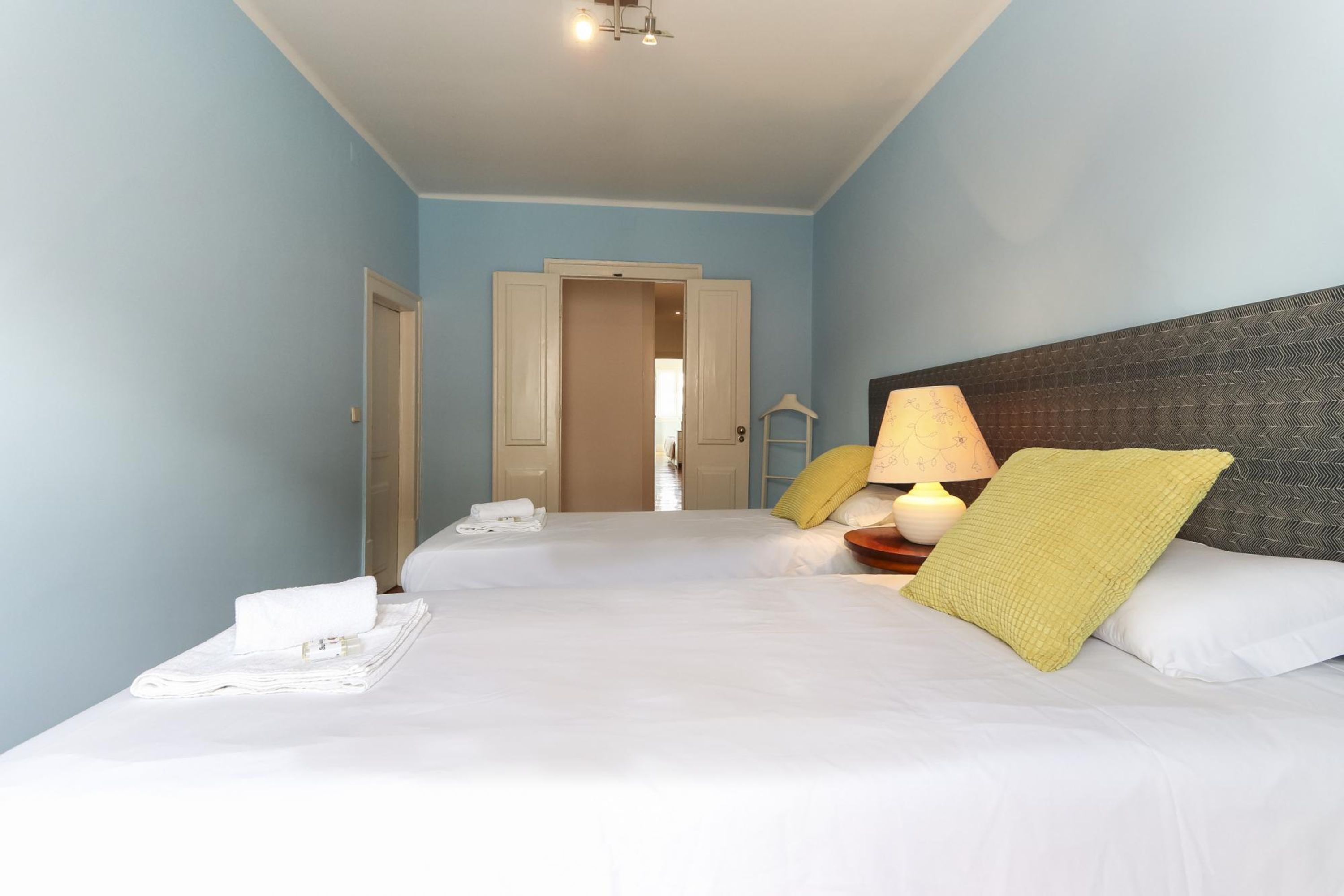 A hotel room with a bed, a nightstand, a lamp, and a door leading to another room.