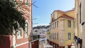 A narrow street with buildings on either side, outdoor seating, and a view of a hill with buildings in the distance. - Chiado Views by Homing (Lisbon)