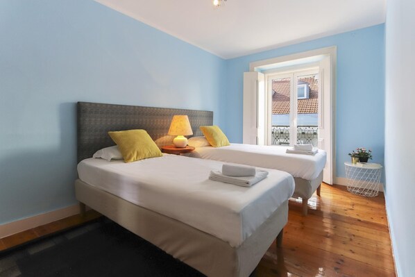A hotel room with two beds, a window with a view of a tiled roof, and a small side table with a lamp and a plant.