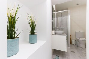 Ein modernes Badezimmer mit Duschkabine aus Glas, weißem Waschbecken und Toilette. Auf einem weißen Regal stehen zwei Blumentöpfe.