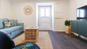 Un salon comprenant un canapé, une petite caisse en bois, un tapis tissé, une armoire verte et un téléviseur.