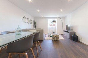Una sala de estar moderna con una mesa de comedor de vidrio, sofás grises, un otoman estampado y un mueble para televisión.