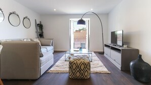 A modern living room with a sofa, ottoman, glass coffee table, and a television on a cabinet. - Benfica Apartments III by Homing (Lisbon)