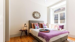 Un dormitorio con cama, mesita de noche con lámpara, ventana con vista a edificios y un espejo decorativo en la pared.